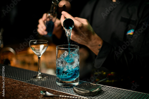 Professional male bartender pouring a blue alcoholic drink from the jigger to a measuring cup holding a glass bottle