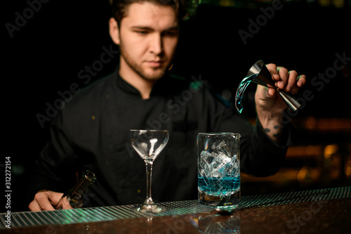 Professional male bartender pouring a blue alcoholic drink from the jigger to a measuring cup