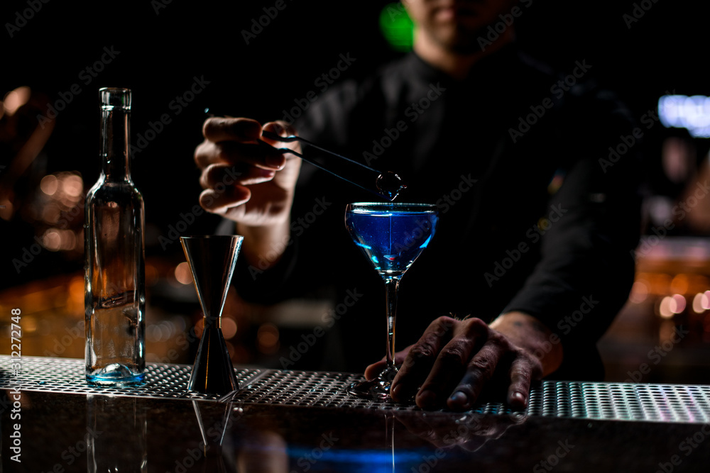 Bartender throwing to the blue alcoholic drink one berry with tweezers