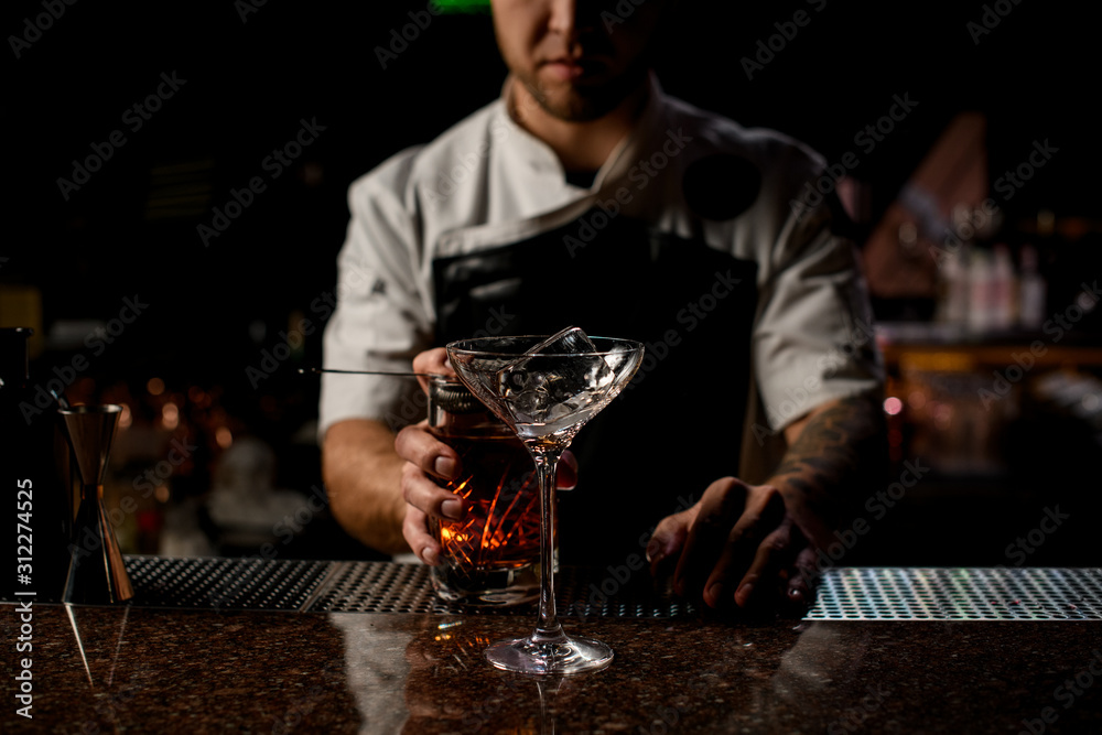 Professional male bartender pouring a brown alcoholic cocktail from the measuring cup to the glass