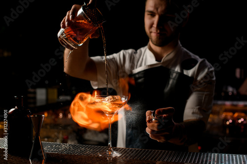Professional male bartender pouring a brown alcoholic cocktail from the measuring cup to the glass burning the fire