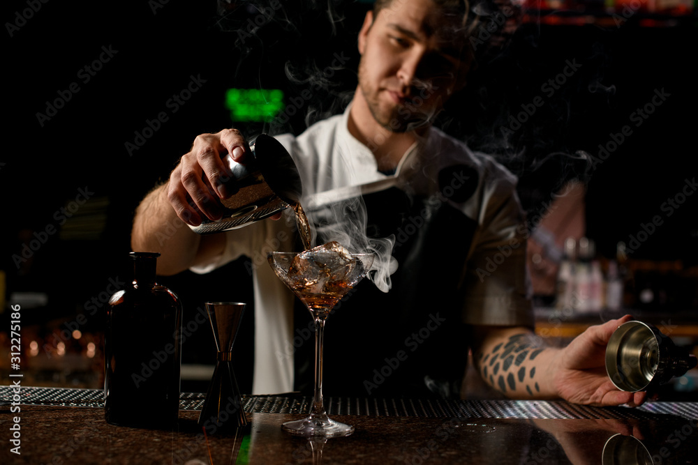 Male bartender pouring a brown alcoholic drink from the steel shaker to the glass with the steam