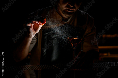 Professional bartender spraying on the brown alcoholic drink with a orange zest juice in the dark