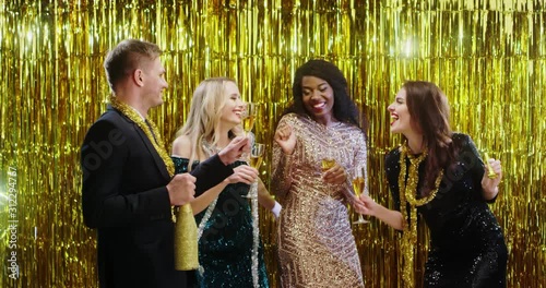 Cheerful young multiethnic company of friends - guy and girls in fancy outfits drinking chamapagne on the New Year Eve and laughing. photo