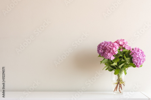 Fototapeta Naklejka Na Ścianę i Meble -  Pink hydrangea flowers with green leaves in glass vase on white side board against neutral wall background