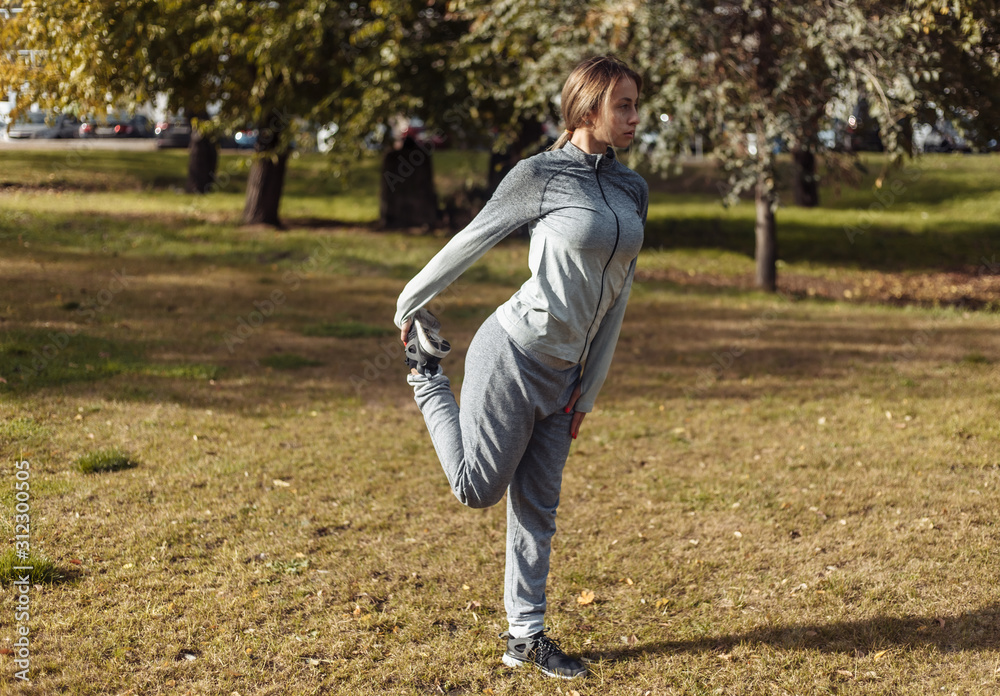 Young sport woman in a sports suit is warming up in the autumn park. Stretching legs before training. Fitness training outdoors