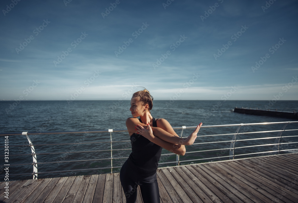 Slim fit woman in sportswear doing stretching hand exercise on the beach at bright sunny day