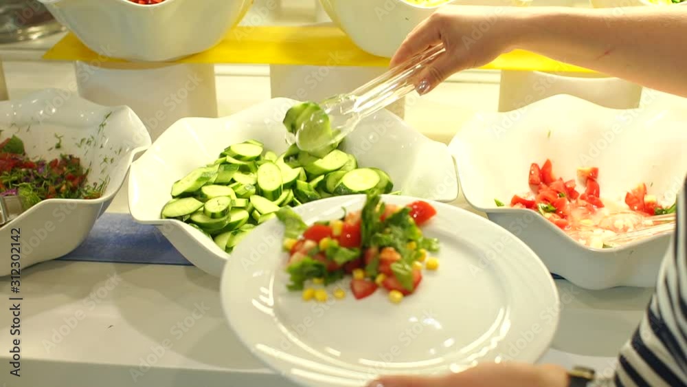 Close-up of a woman puts a vegetable salad in a plate with tongs. Self-service restaurant in the hotel. Woman puts the food on her plate from the buffet at the hotel. Breakfast buffet at a hotel.