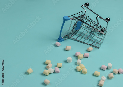 Lot of mini marshmallows and shopping trolley on a blue background. Pastel color trend