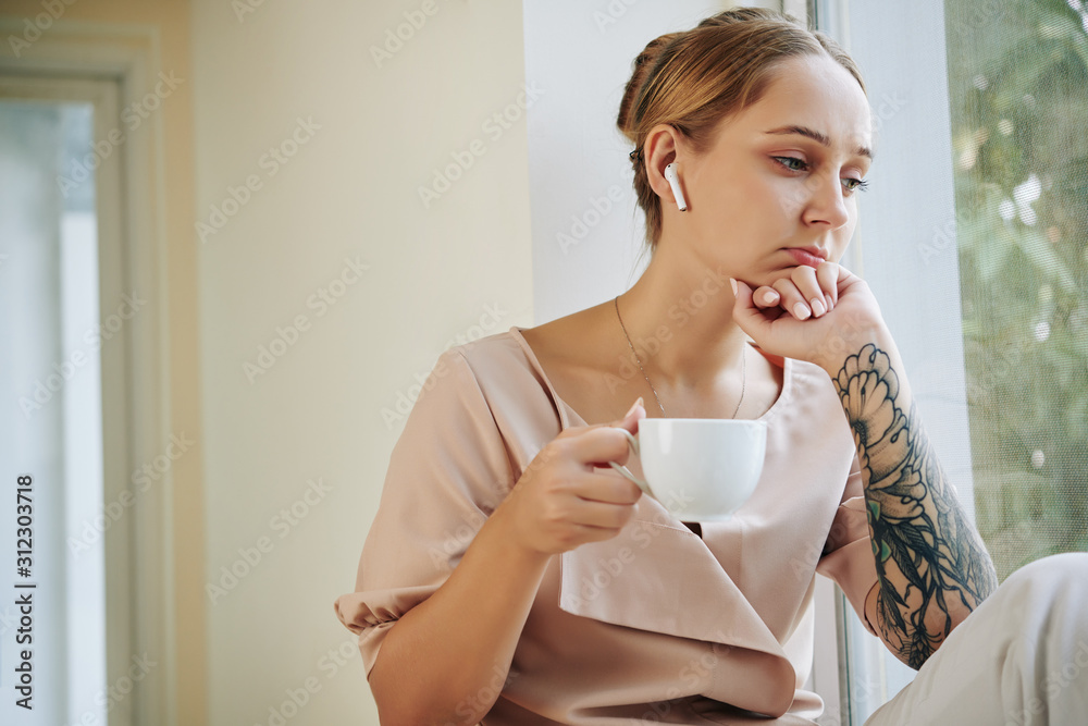 Young woman listening to serious audiobook in her earbuds when sitting on window sill and drinking cup of tea