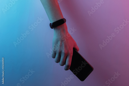 Female hand with smart bracelet and smartphone. Modern gadgets. Creative pop art pink blue neon color. Trendy gradient illumination photo