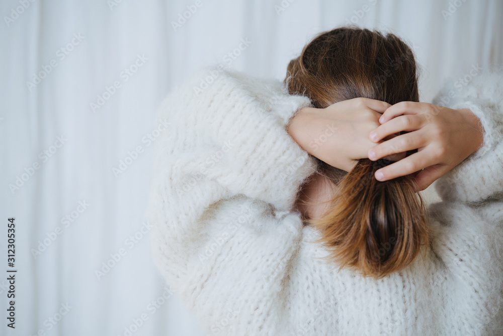 Brunette little girl in white fluffy knitted sweater putting her hair in a pony tail. At home, in front of a curtain. From behind. Brunette little girl in white fluffy knitted sweater putting her hair in a pony tail. At home, in front of a curtain. From behind.