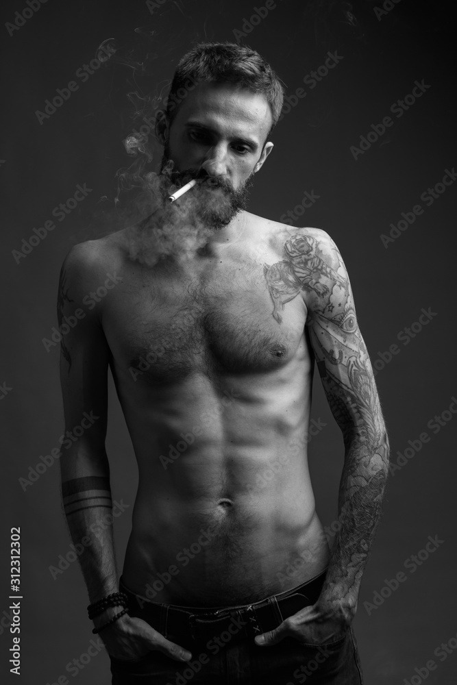 A charismatic young man with a naked torso with tattoos and a smoking cigarette. Black and white image