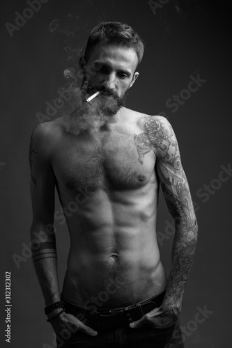 A charismatic young man with a naked torso with tattoos and a smoking cigarette. Black and white image