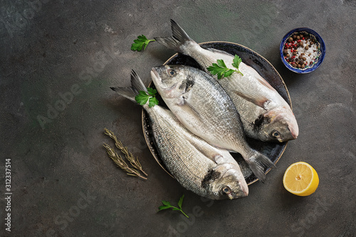 Fresh raw dorado fish in a plate with lemon, spice and parsley on a dark paint background. Top view, flat lay.