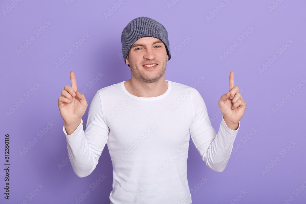 Studio shot of young caucasian man wears cap and white casual shirt, stands smiling isolated purple background, points up with fingers and raised arms. Copy space for advertisment or promotional text.