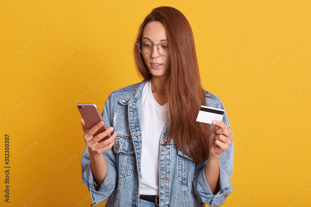Φωτογραφία Portrait of concentrated young woman wearing white casual shirt and denim jacket, using mobile phone and holding credit bank card, posing isolated over yellow background