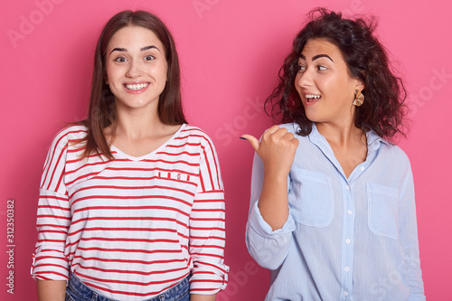 Front view portrait of charming astonish woman standing and pointing with her thumb to her smiling female wearing striped casual shirt, brunette girls posing isolated over pink studio background.