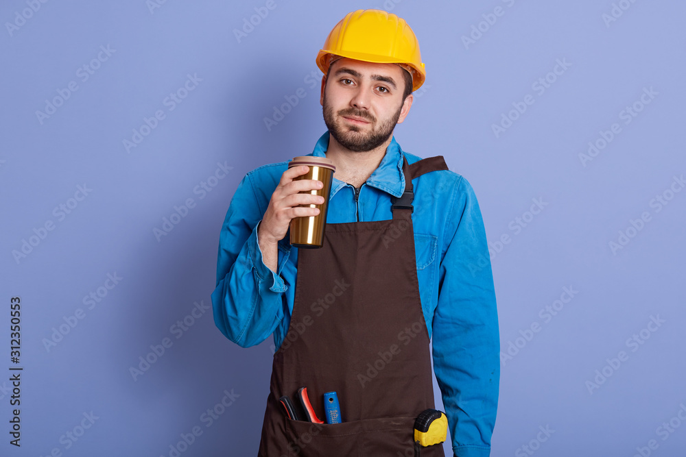 Picture of bearded handsome repairman with thermo mug in hand, wearing hard hat and special uniform, enjoying hot coffee or tea, looking directly at camera and posing isolated over blue background.