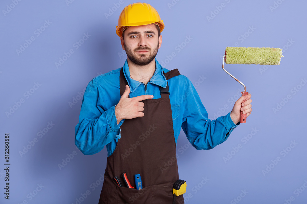 Close up portrait of repairman wearing yellow protective hat, apron and uniform, holding paint roller for painting wall and pointing at it with his index finger, craftsman isolated on blue background.