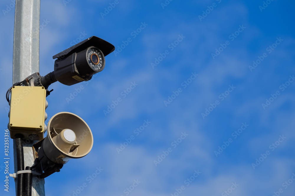 CCTV camera and loudspeaker alarm on a pole against a blue sky. CCTV camera and loudspeaker alarm on a pole against a blue sky.
