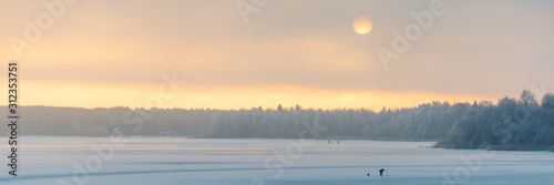 Fantastic winter landscape. Shore of a frozen river covered with snow. The disk of the sun shines through fog and clouds. An orange beautiful dawn dawns upon fishermen in the distance. Web banner.