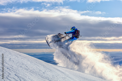 a snowmobile rider jumps in a mountain valley at dawn. sports snow bike with snow splashes and snow trail. bright snowmobile and suit without brands. snowmobilers sports riding. stock photo photo