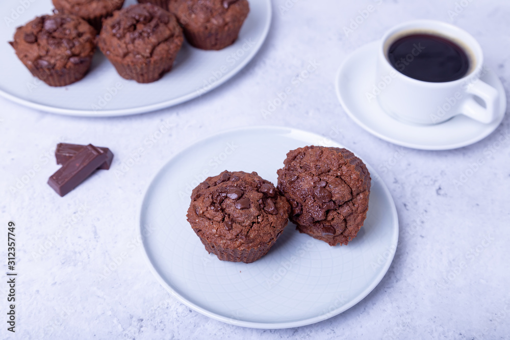 Chocolate muffins on a white plate. Homemade baking. In the background is a cup of coffee. White background. Selective focus, close up. Chocolate muffins on a white plate. Homemade baking. In the background is a cup of coffee. White background. Selective focus, close up.