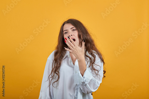 Tired caucasian young woman in fashion white shirt covering mouth with hand, yawning isolated on orange background in studio. People sincere emotions, lifestyle concept.