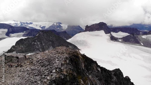 Aerial, reverse, drone shot, of two mountaineers, on the Galdhopiggen mountain, overlooking the Svellsnosbrean glacier, on a overcast day, in Jotunheimen National park, in Norway photo