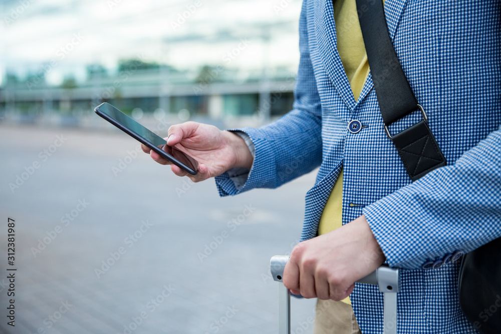 Stylish man standing at airport with smartphone and suitcase, browsing, texting, using mobile app. Businessman traveling. Close-up
