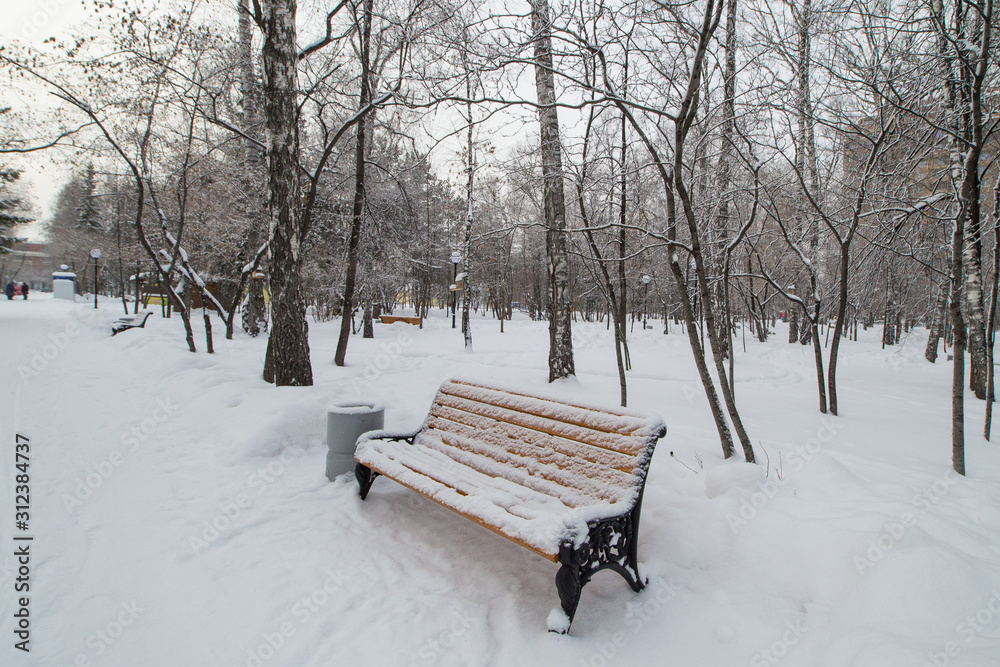 A bench covered with snow in a city Park in winter, with people in the background