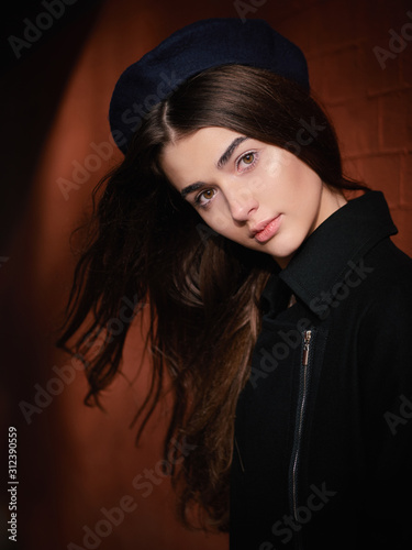 Mood contrasty portrait young elegant french style brown haired woman in beret and black leather jacket liten sun spot cinematic stock colour grading city street red bricks arc background