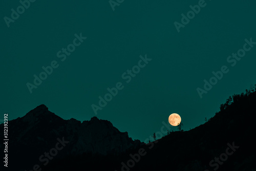 Minimalist spooky night landscape background with mountainous range silhouette and the Moon at moonrise time. Scary Happy Halloween scene on dark teal blue sky and black backdrop.