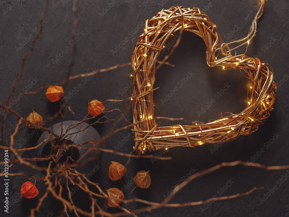 A golden wicker heart and a luminous garland are on a dark background. Decorating with dried flowers and branches. Composition for Valentine's Day. Flat lay, copy space.
