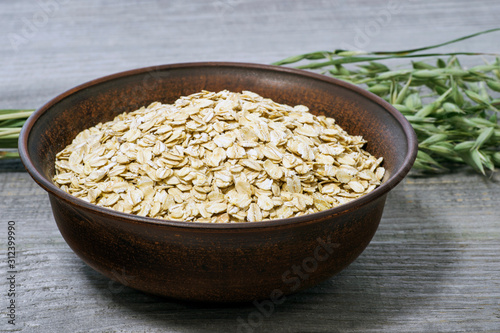 Oatmeal flakes in a deep ceramic brown dark bowl close-up against green oat ears on an old faded gray wooden table.