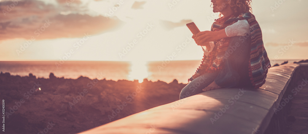 Trendy woman enjoying the phone connection during a coloured beautiful sunset on the coast - outdoor leisure activity for traveler modern people - obrazy, fototapety, plakaty 
