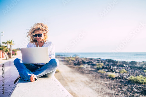 Beautiful caucasian adult woman using modern technology laptop computer in outdoor near the beach and ocean - work everywhere for digital nomad free people concept