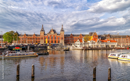 Panoramic view of Central Train Station in Amsterdam, architecture monument built in 1889, city's main public transportation hub. Canal with ferry boats in the foreground. Amsterdam, Netherlands.