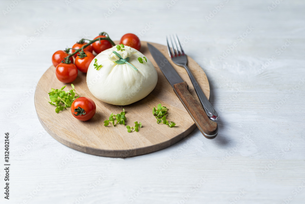 Italian cheese Burrata lies on a round wooden board surrounded by cherry tomatoes and garnished with microgreens. Near cutlery knife and fork. Food on white wooden table. Italian cheese Burrata lies on a round wooden board surrounded by cherry tomatoes and garnished with microgreens. Near cutlery knife and fork. Food on white wooden table.