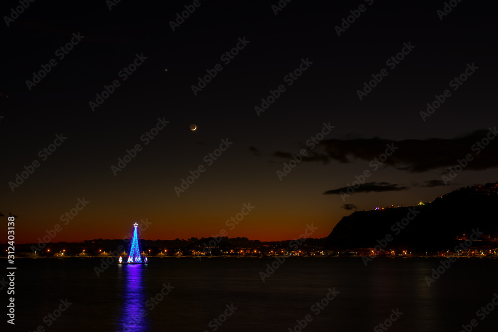 Bacoli Naples, December 27, 2019. An illuminated tree in the middle of the lake with a slice of moon in the background.