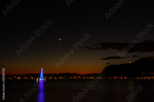 Bacoli Naples, December 27, 2019. An illuminated tree in the middle of the lake with a slice of moon in the background.