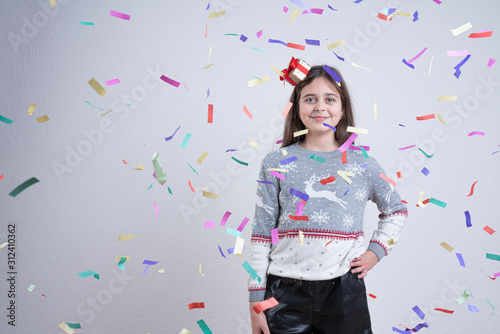 Happy brunette girl in grey santa sweater and hat stands among multi-colored confetti holds her hand on her waist and looking at the camera, isolated over grey background, Happy New Year and Merry