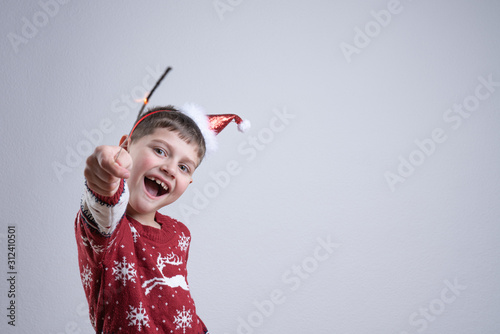 Blissful smiling child boy in red santa sweater and hat holds in his hand sparkler, isolated over grey background, Merry Christmas and Happy New Year concept