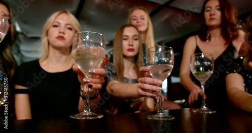Seven fateful girls in bright shiny dresses dance, hang out and have fun at a party in a bar-club. They have glasses of alcoholic cocktails in their hands and are sitting at the table.