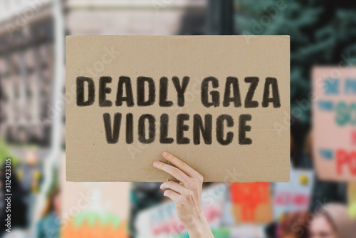 The phrase " Deadly gaza violence " on a banner in men's hand. Human holds a cardboard with an inscription. Kills. Refugee. War. Conflict. Safety. Palestine. Protest