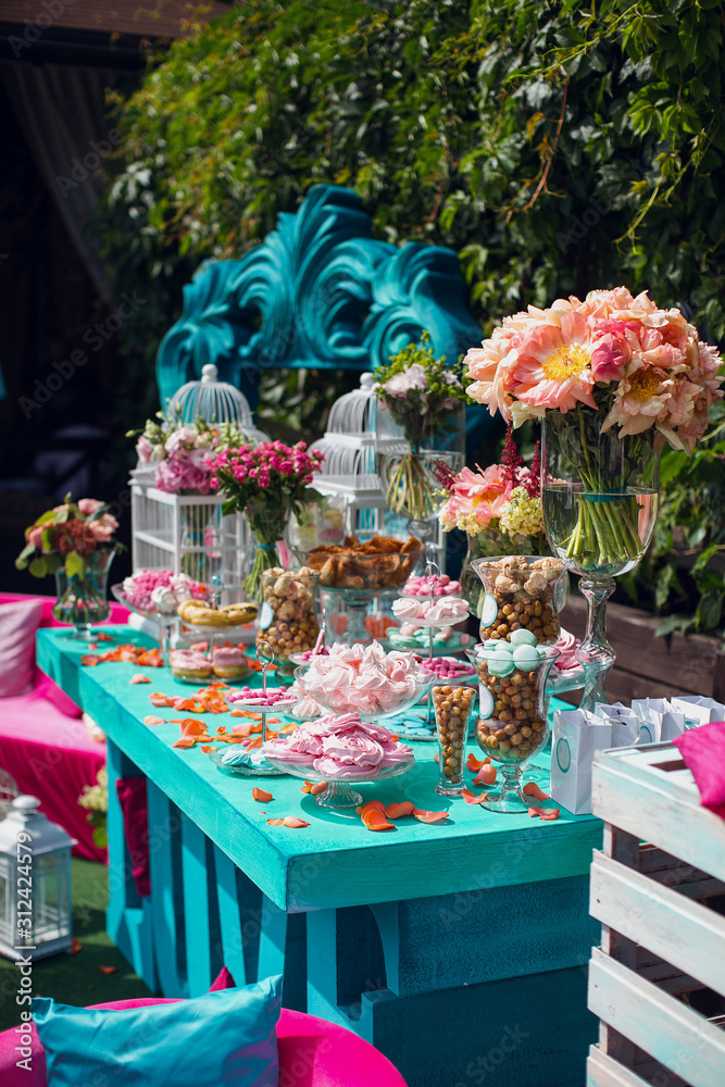 Decorative sweet wedding table in bright colors of blue, turquoise and pink. Decorated with lanterns, pillows, upholstered furniture, watches and flowers. Candy bar with macaroons, meringues, cookies.