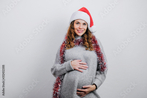 Pregnant girl wearing Santa hat smiling on grey background. Expecting a child. Winter pregnancy. © meteoritka