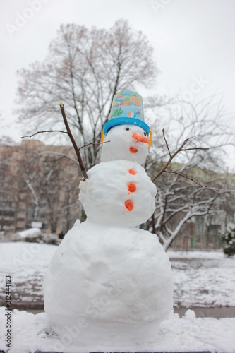 Cheerful snowman with carrot nose and blue bucket on his head in winter snowy park. Bottom view. Snowman in the snow © Juver