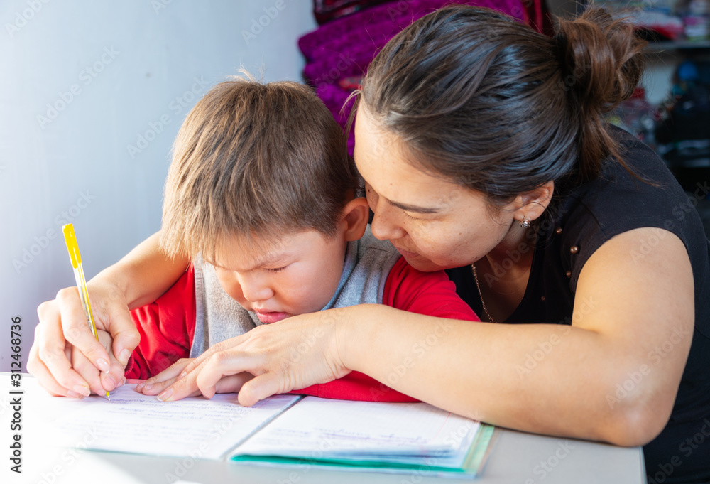 Concentrated child doing homework at home with mom. The boy sits and looks into books and notebooks. Concentrated child doing homework at home with mom. The boy sits and looks into books and notebooks.